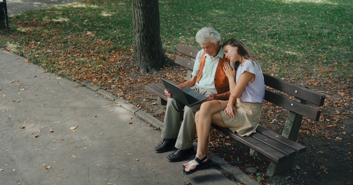 Ein älterer Mann sitzt mit einer Frau auf einer Bank und blickt auf einen geöffneten Laptop.